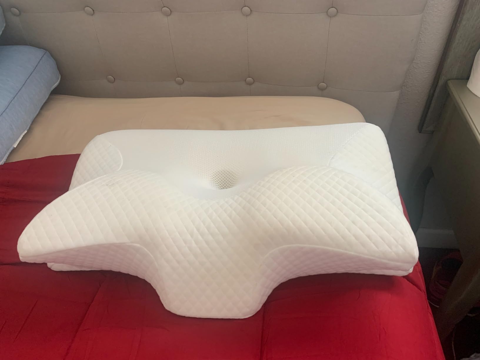A white HOMCA Cervical Pillow rests on a bed alongside a red pillow, showcasing its design in a cozy setting.