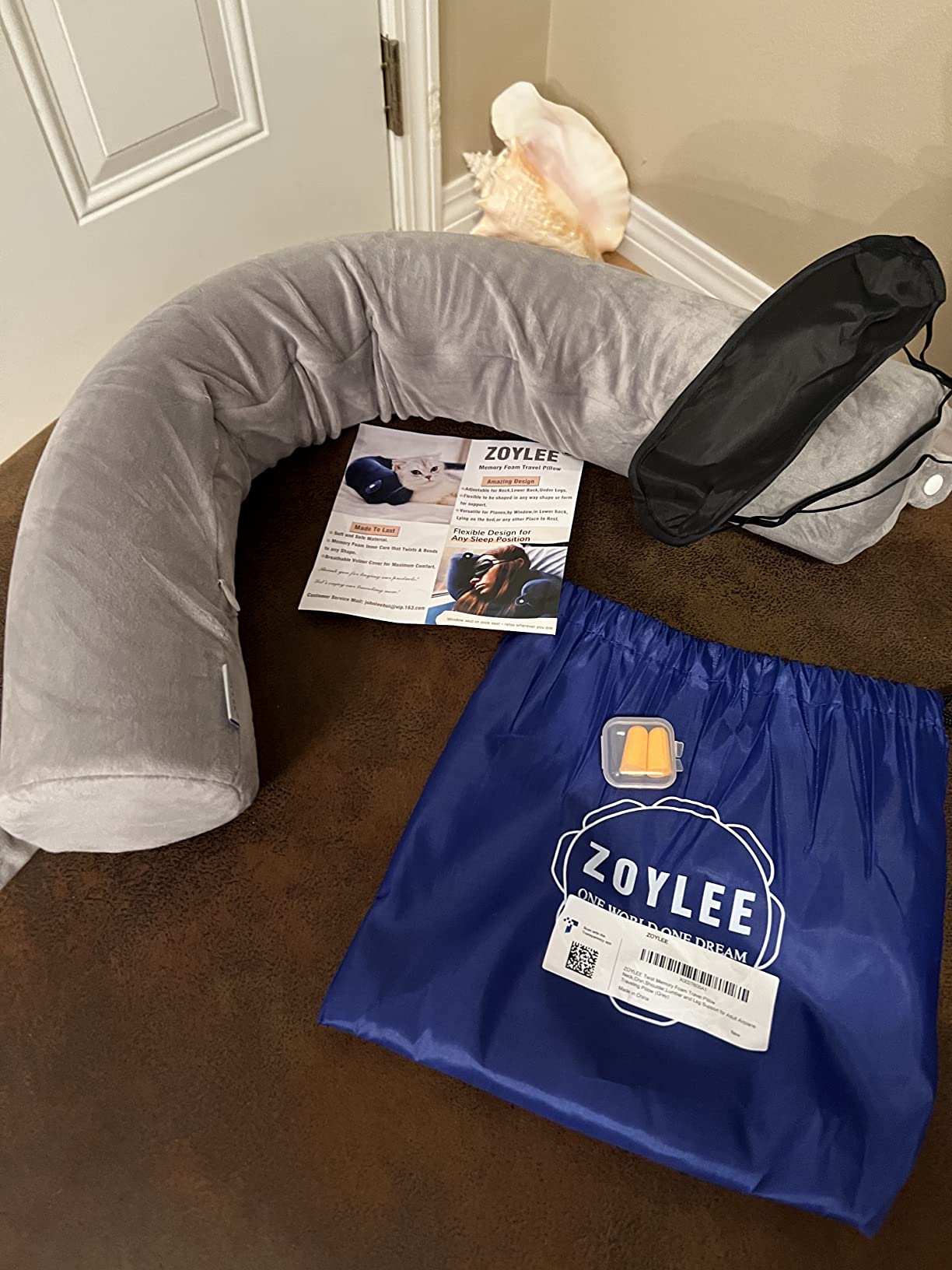 A ZOYLEE Twist Memory Foam Travel Pillow rests on the floor alongside a bag and a magazine, perfect for a casual review.